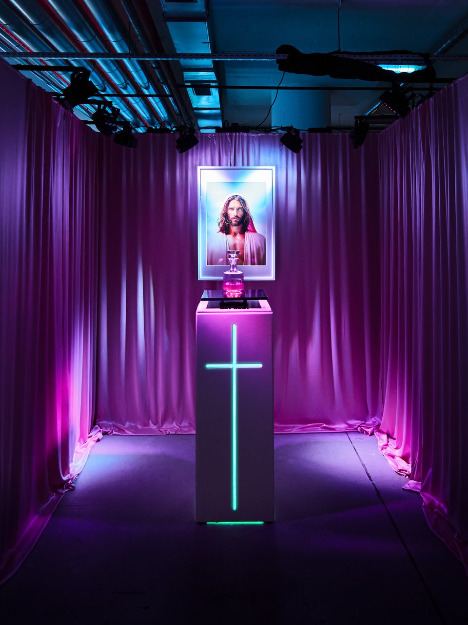 Die echte Installation in Fulda: Porträt im weißen Rahmen, Karaffe auf Podest mit Neon-Kreuz, rosa Vorhänge